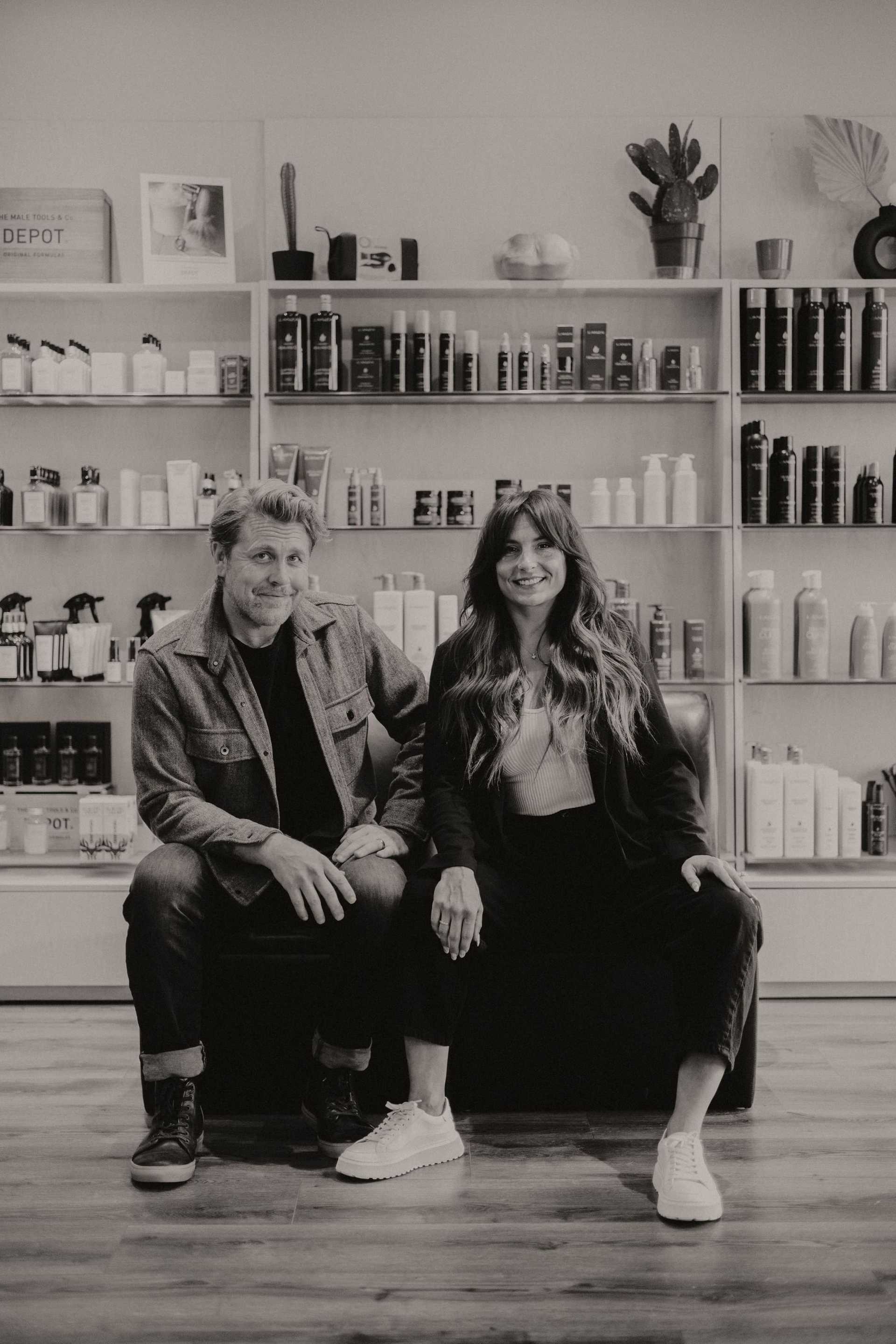 Man and woman smiling, sitting in salon with shelves of hair products behind them.